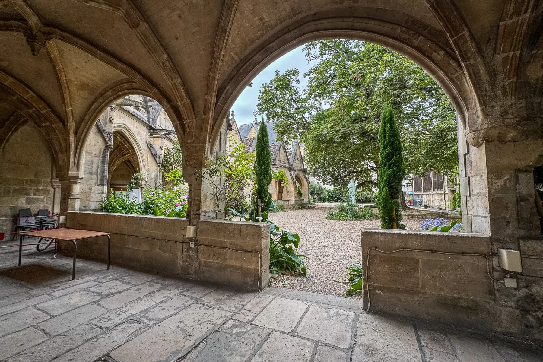 View through gothic stone arches into a gravel courtyard with trees, a statue, and historic buildings under a cloudy sky.