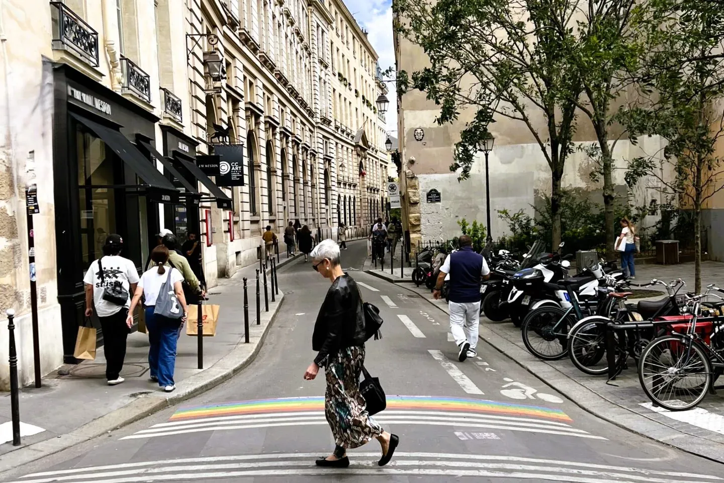 People walk on a Parisian street with a rainbow-colored crosswalk, flanked by classic buildings, parked motorcycles and bi...