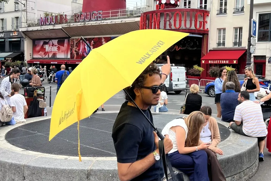 A man in sunglasses holding a yellow Walkative umbrella, pointing, with the red Moulin Rouge building in Paris visible in ...
