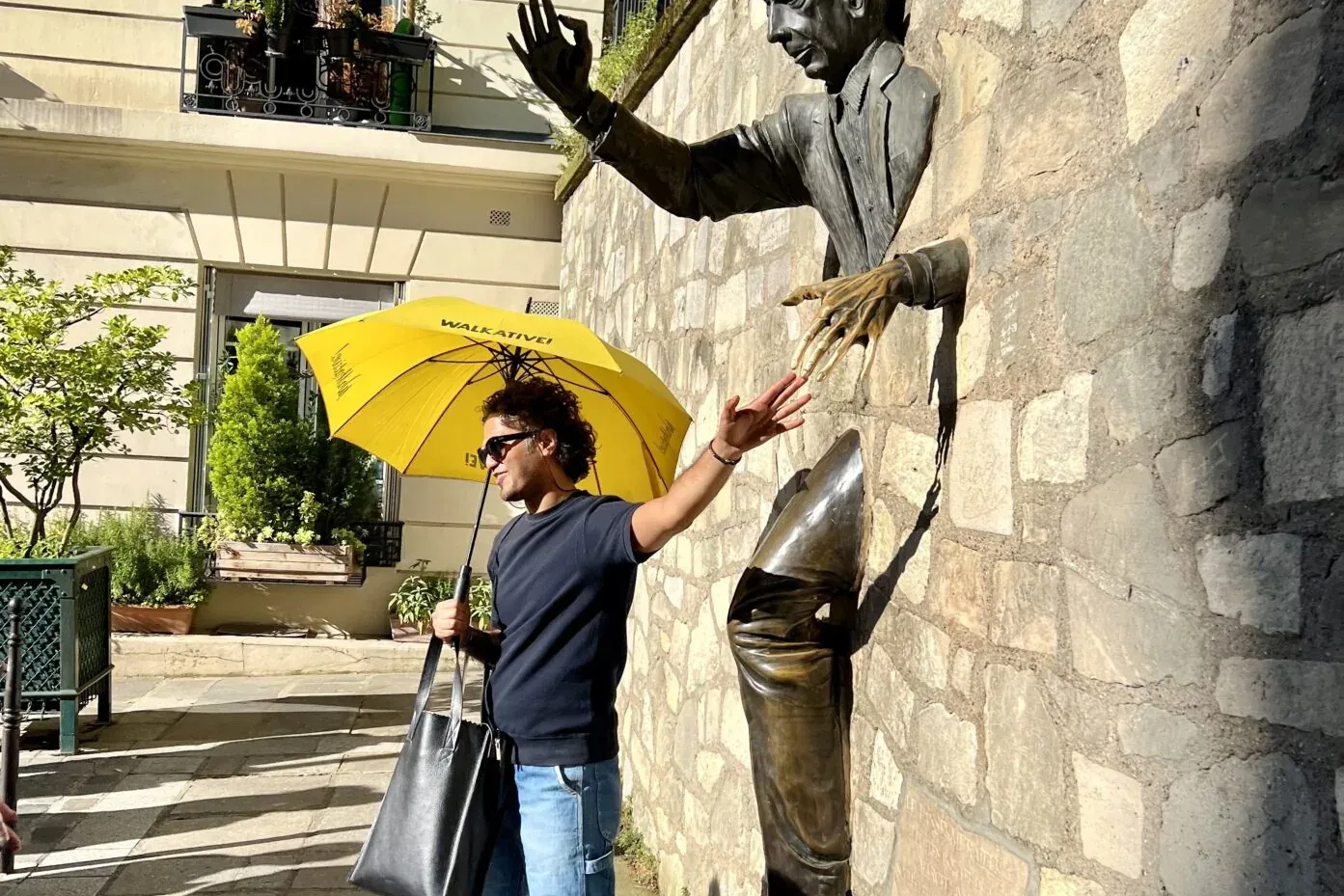 Man with a yellow Walkative umbrella interacting with a bronze sculpture of a man emerging from a stone wall in sunlight.