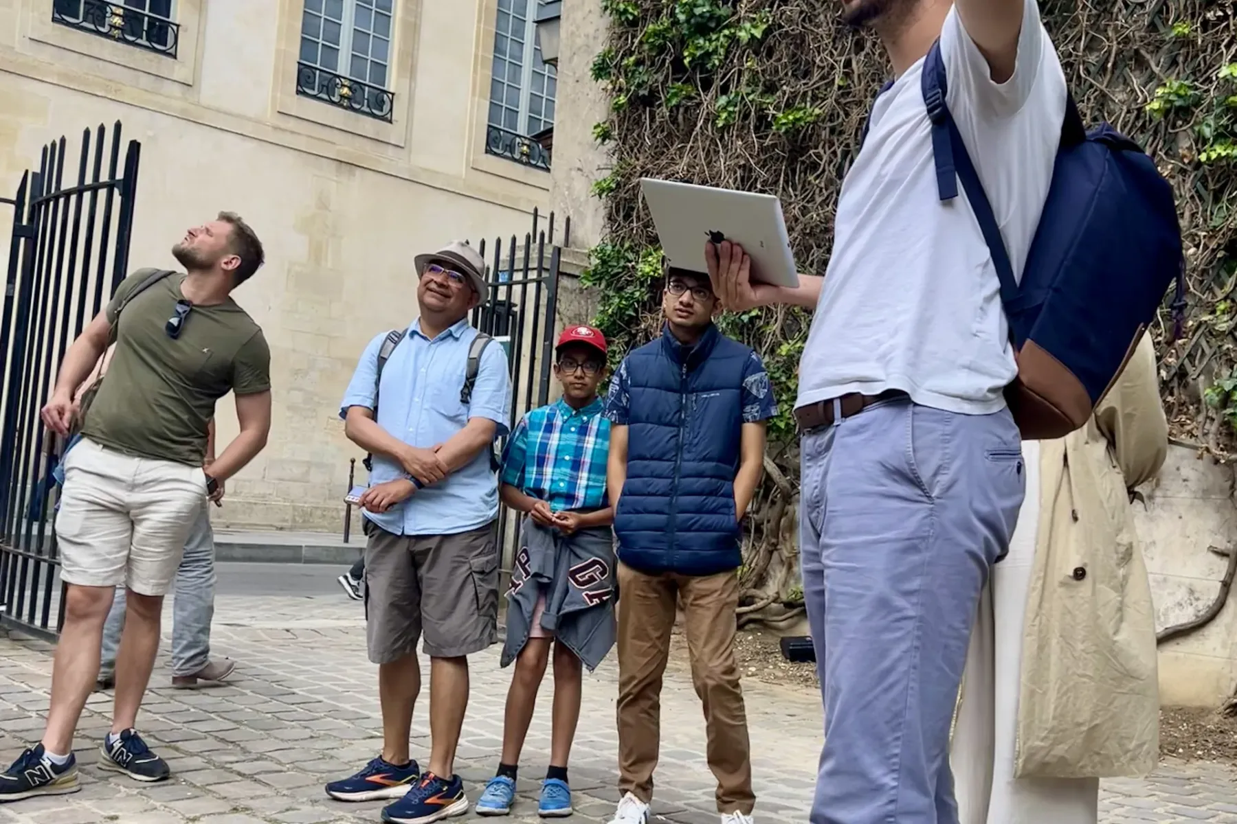 Tour guide holding a tablet and gesturing to a group of five tourists on a cobblestone street in front of a stone building...