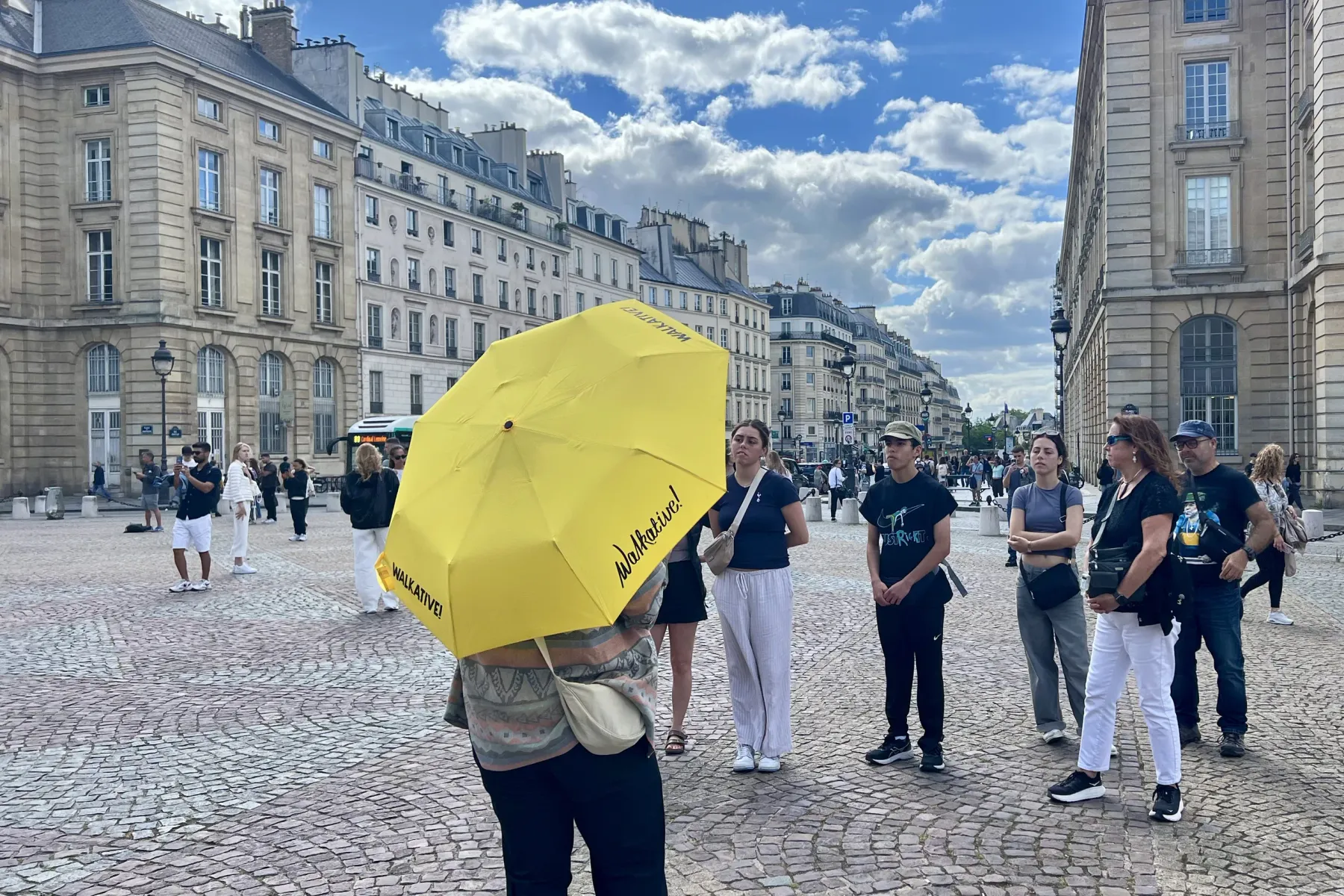 Back of a person holding a yellow Walkative umbrella on a cobblestone street. Other people and Parisian buildings are in t...
