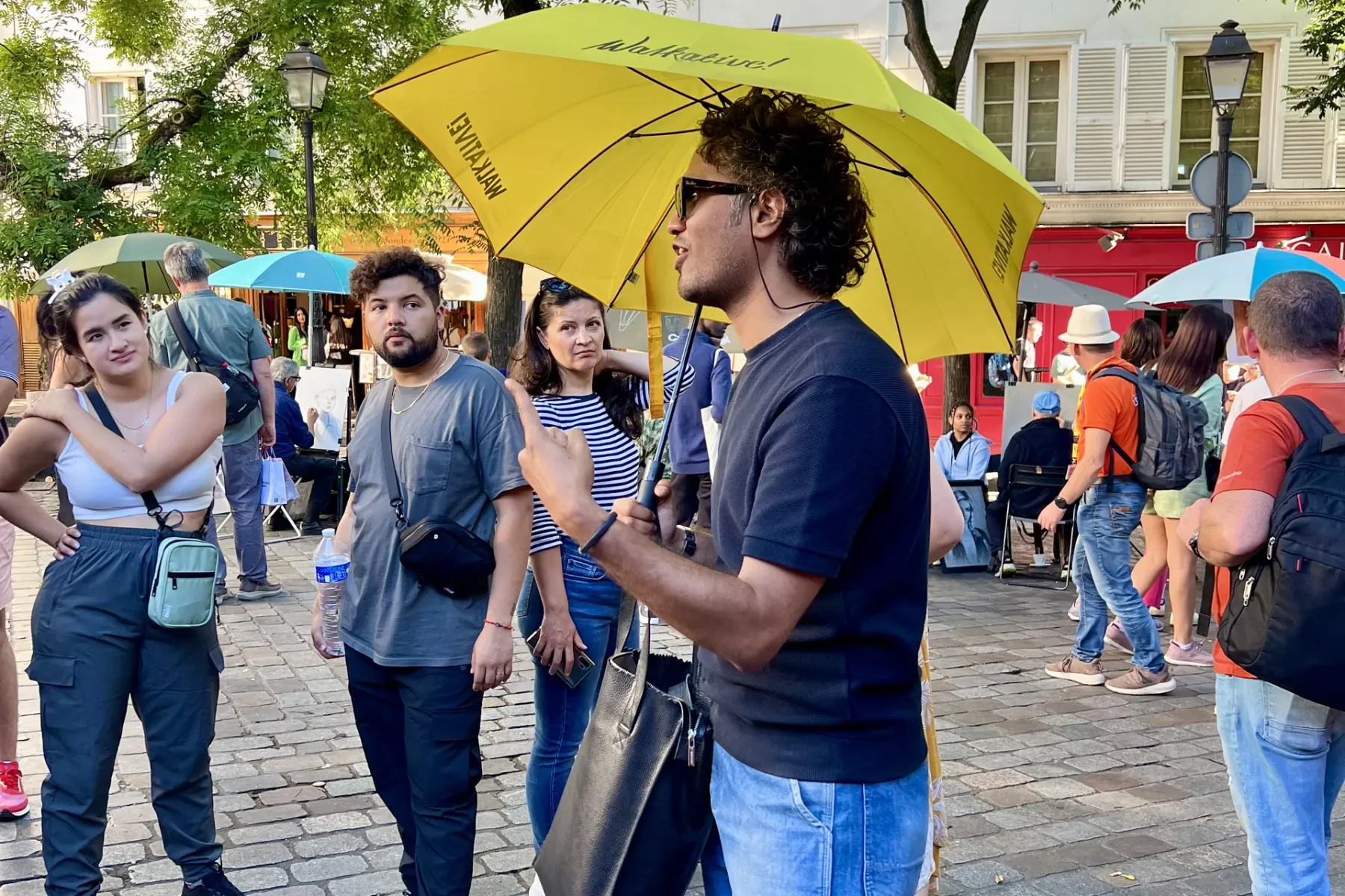 A tour guide under a yellow Walkative umbrella speaks to a small group of tourists on a cobblestone street in Paris.