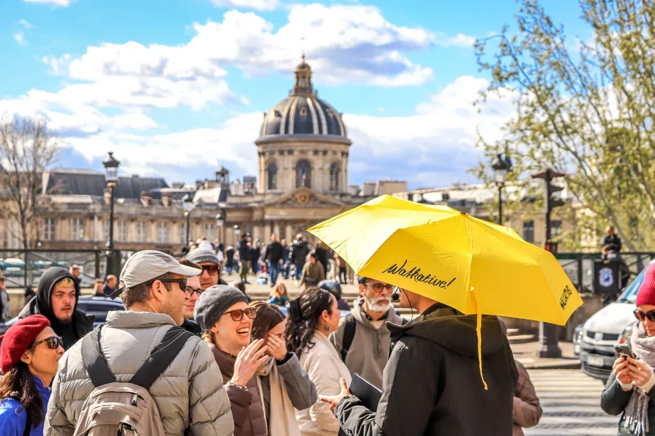 A group of tourists listens to a guide holding a yellow Walkative umbrella in Paris, with the Institut de France dome in t...