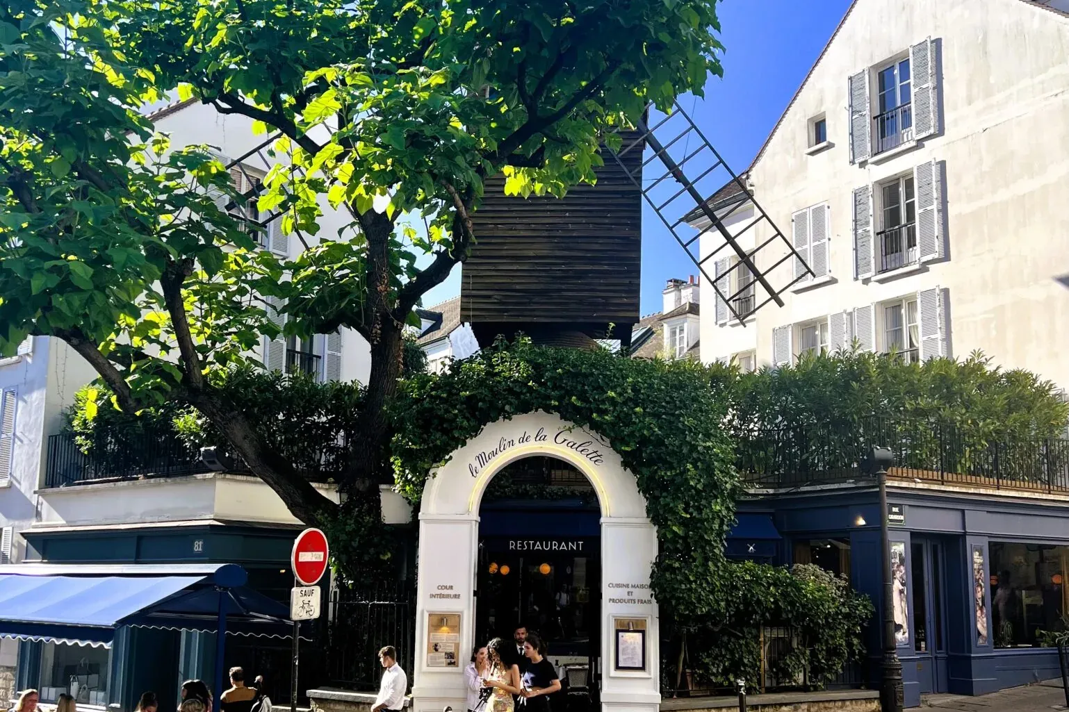 Le Moulin de la Galette restaurant in Paris, with its historic windmill structure, outdoor dining area, green trees, and c...