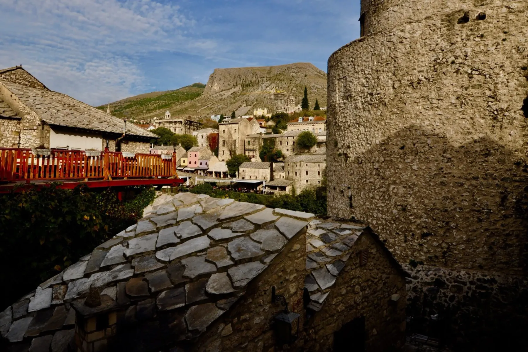 Traditional stone houses with layered roofs, a large round stone tower, and a restaurant terrace in a hillside village, wi...