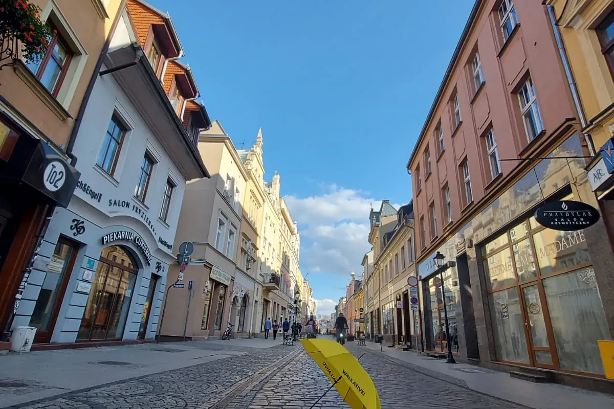 A low-angle view of a cobblestone street in Bydgoszcz lined with historic buildings, featuring a yellow Walkative umbrella...
