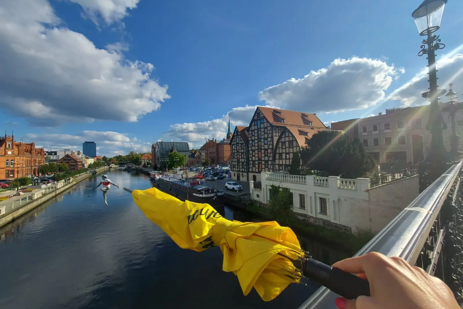 Hand holding a yellow umbrella over the Brda River, with a tightrope walker sculpture, historic buildings, and a blue sky ...