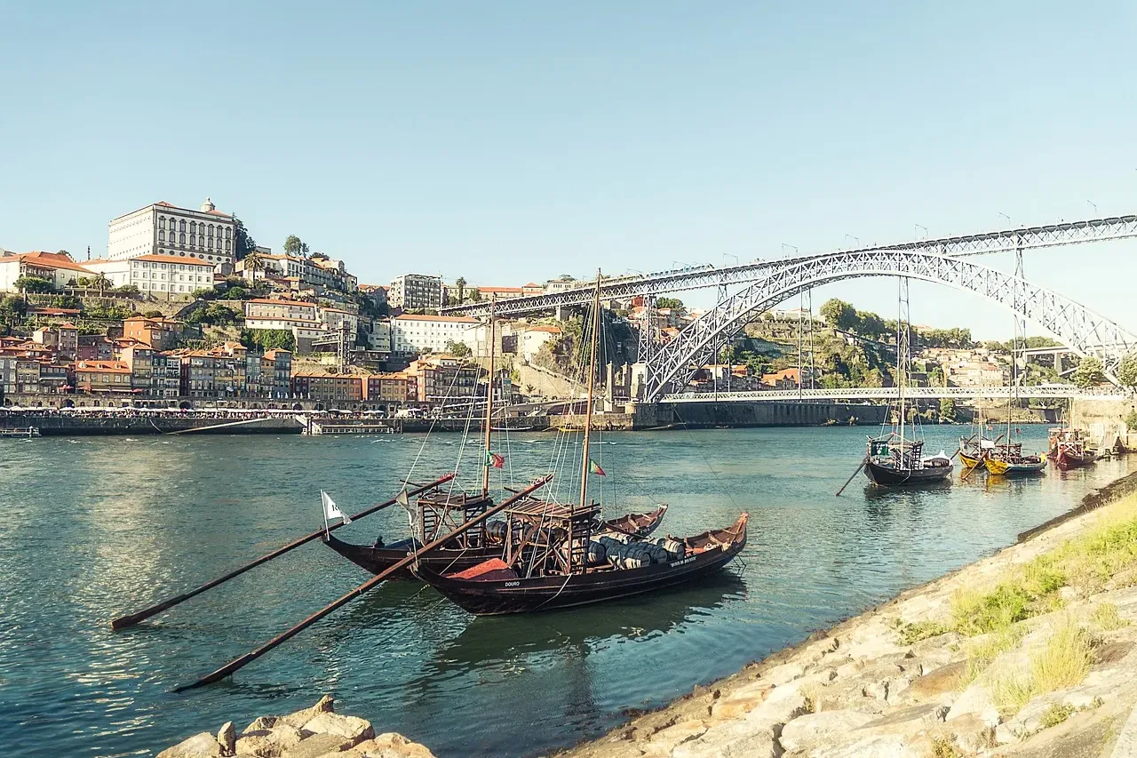 Traditional Rabelo boats float on the Douro River, with Porto's colorful buildings and the Dom Luís I Bridge in the backgr...