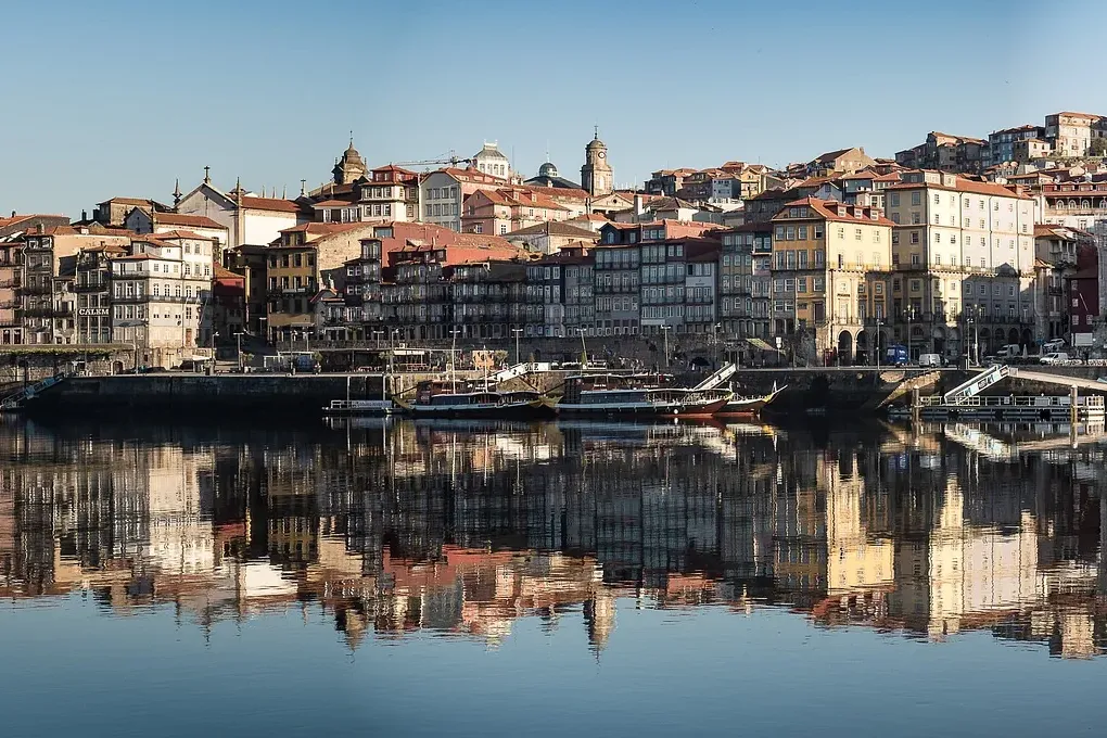 Historic multi-story buildings with red roofs lining a river, reflecting in the calm water. Traditional boats are docked a...