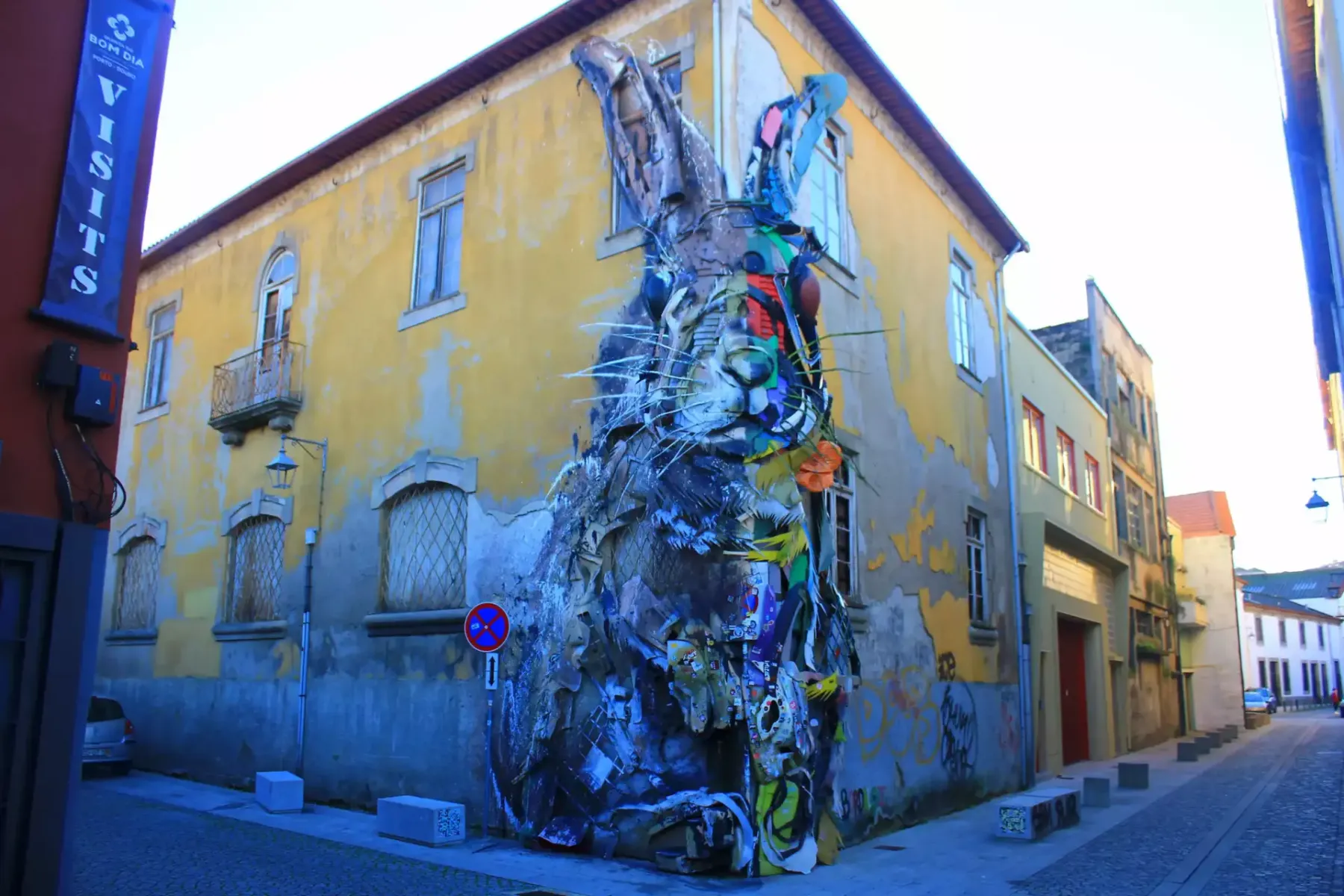 Large rabbit sculpture made from recycled materials attached to a yellow building corner in Porto. Cobblestone street.