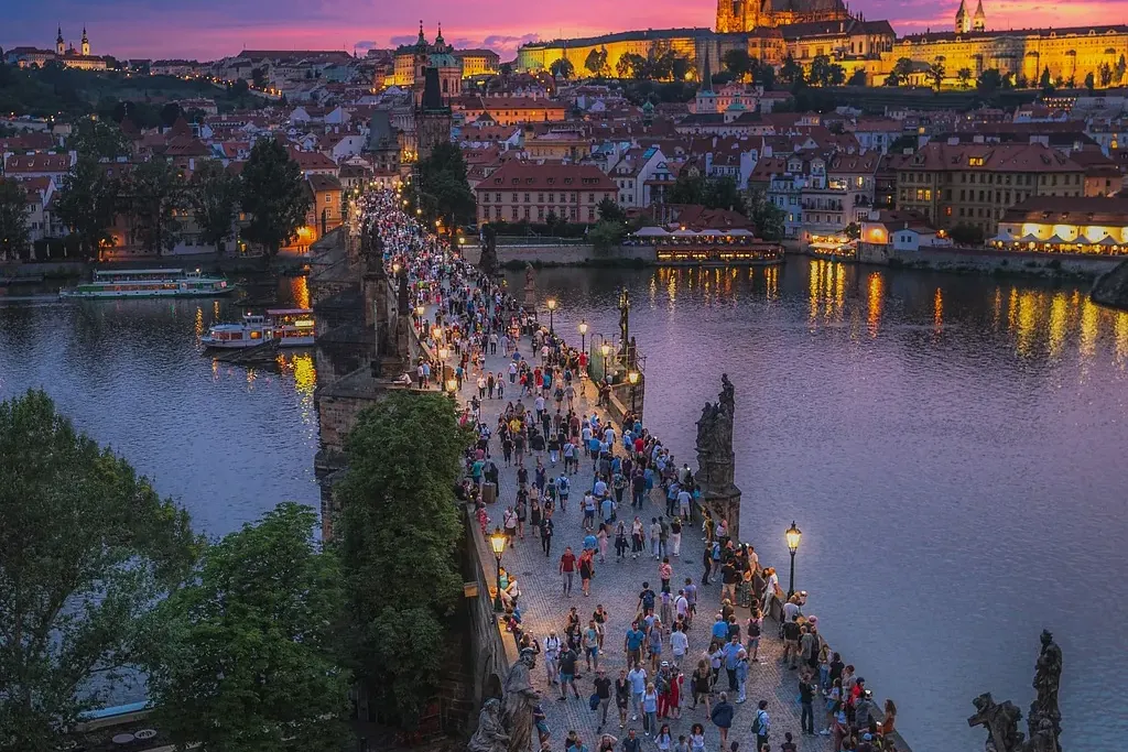 Charles Bridge in Prague bustling with pedestrians at sunset, with Prague Castle illuminated in the background across the ...