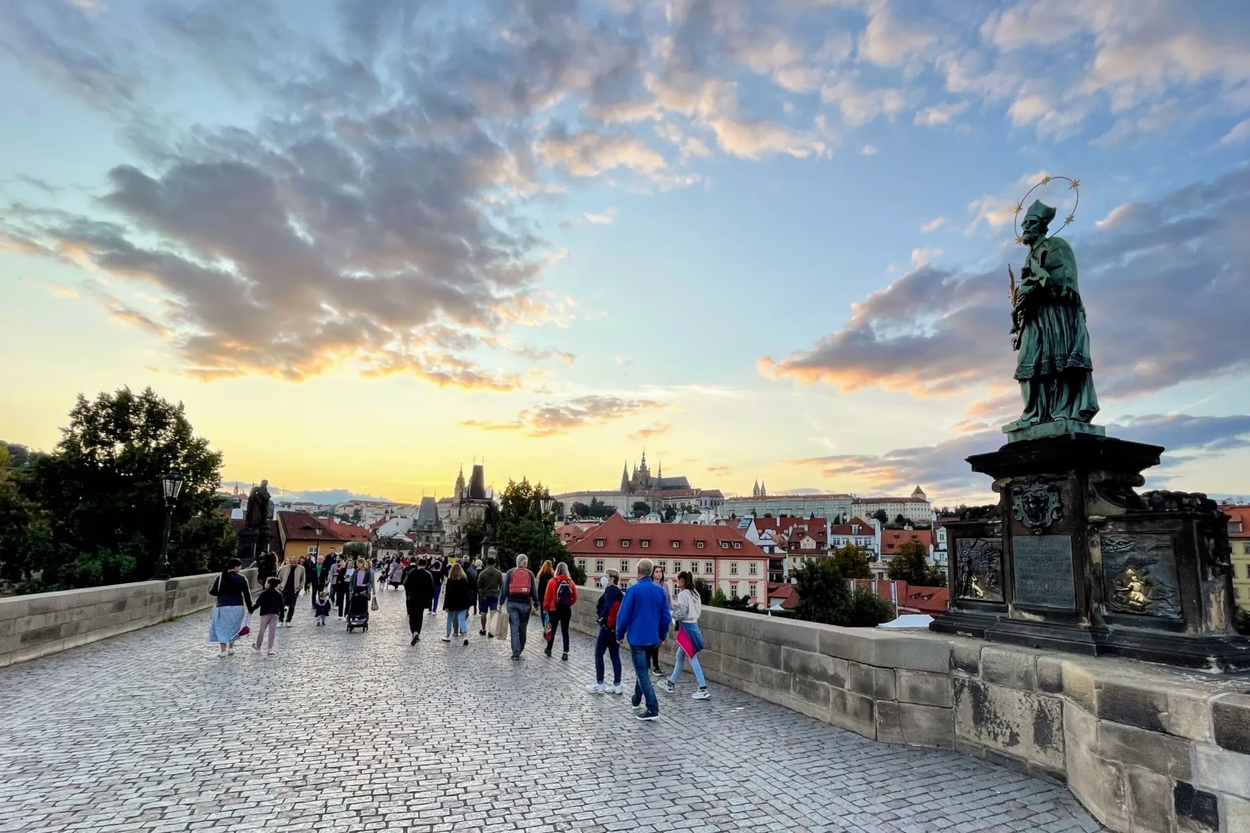 Cobblestone Charles Bridge with people walking at sunset, a statue of St. John of Nepomuk on the right, Prague Castle in b...