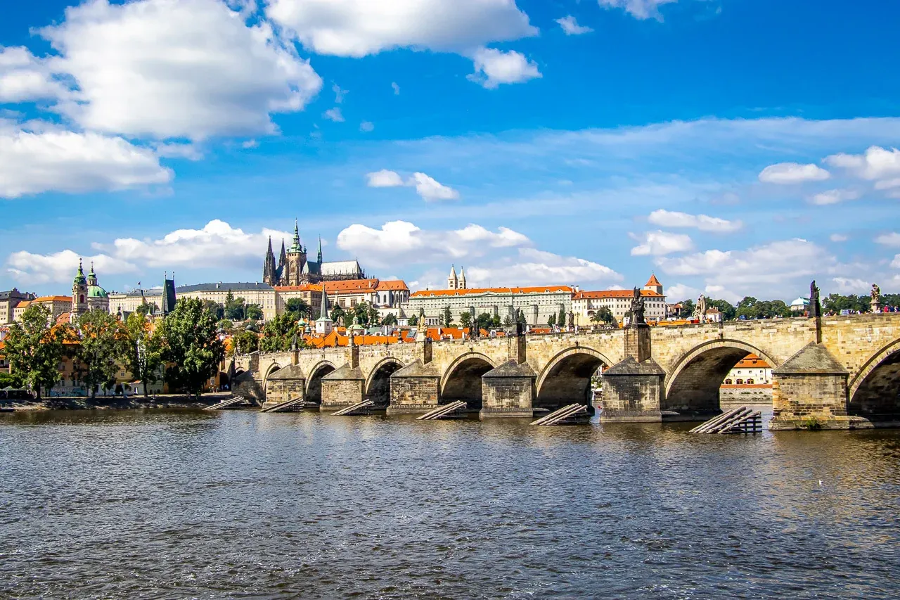 View of Charles Bridge with statues and arches spanning a river, Prague Castle on the distant left bank under a blue sky w...