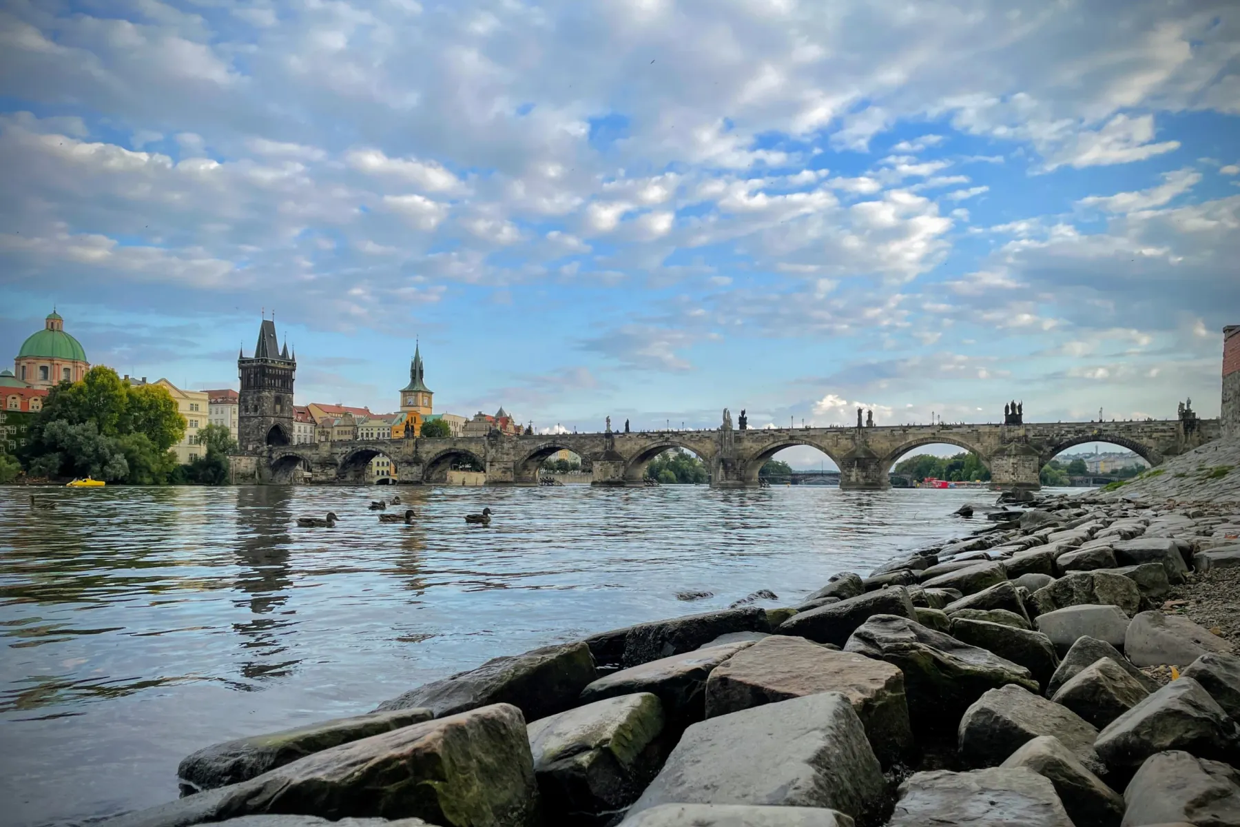 View of Charles Bridge from the Vltava riverbank, with ducks, historic buildings, and a cloudy sky.