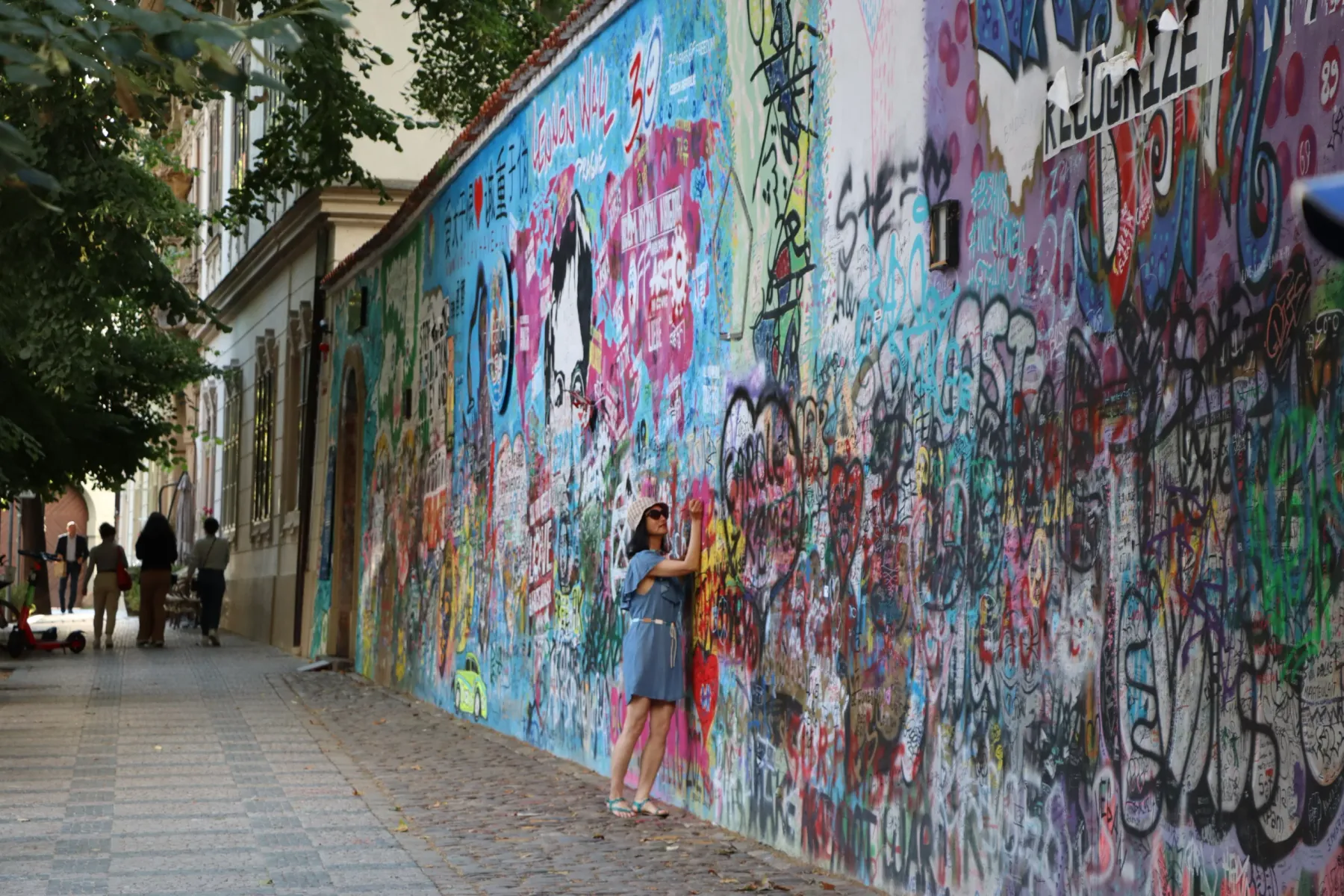 A woman in a blue dress and straw hat stands next to Prague's Lennon Wall, covered in colorful graffiti.