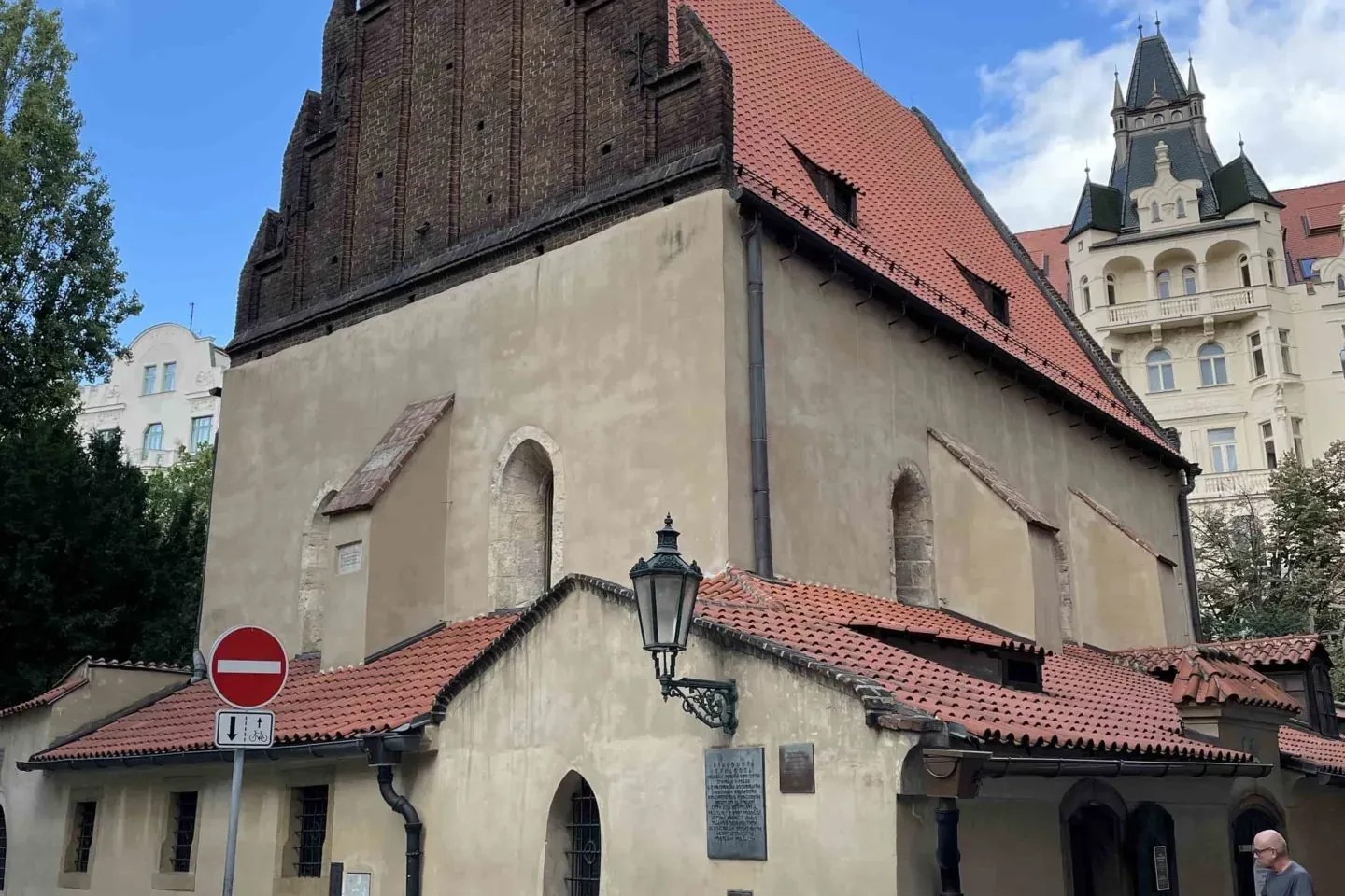 Exterior of the Old-New Synagogue in Prague with its brick gable, terracotta roof, and stucco facade, a person walks on a ...