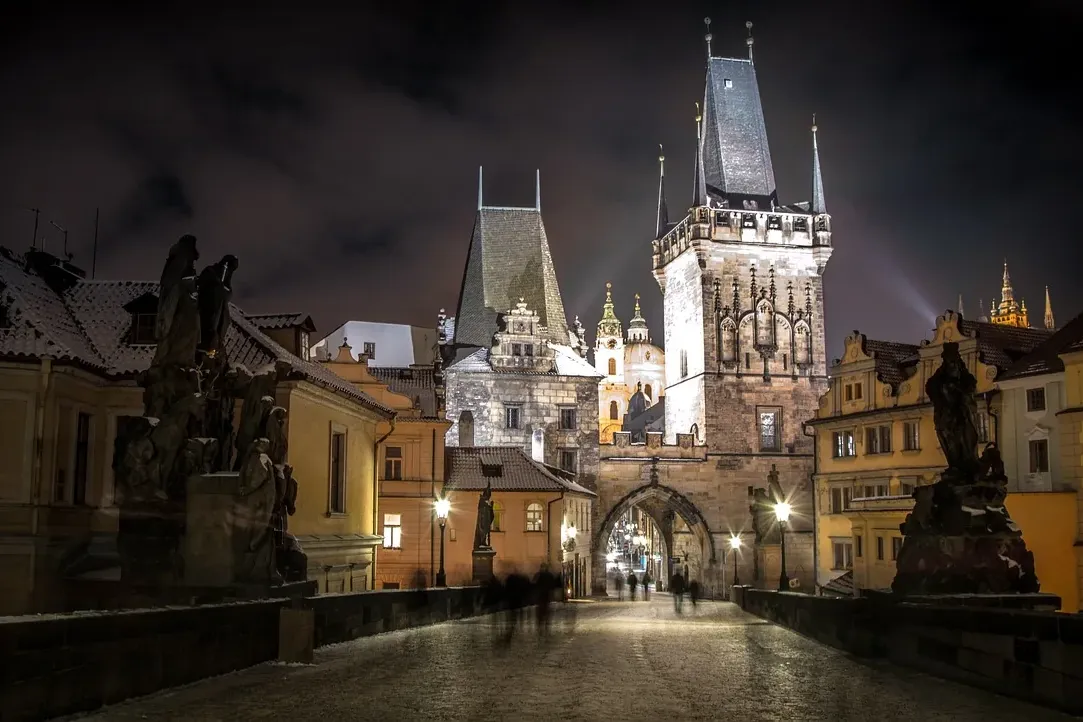Charles Bridge at night, illuminated Old Town Bridge Tower with snow on rooftops and faint figures walking on the bridge.