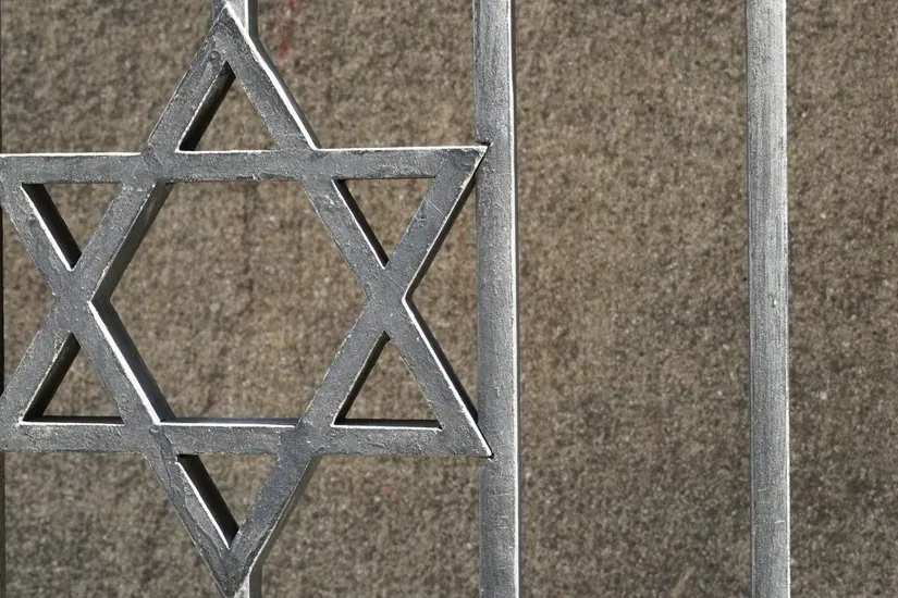 Metal fence with a Star of David symbol in the center, against a textured gray wall.