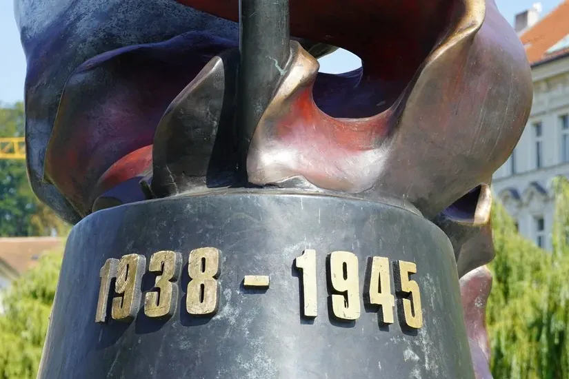 Close-up of a bronze monument base with golden dates '1938 - 1945' inscribed. A church tower and trees are in the blurred ...