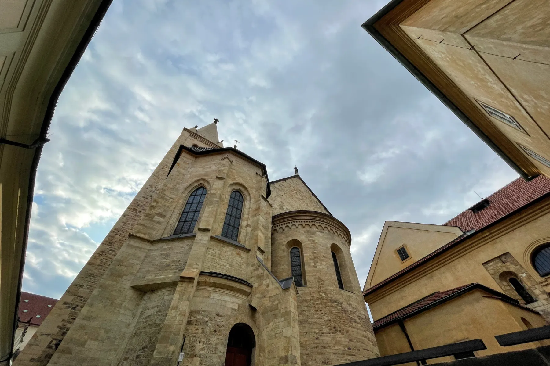 Stone church building with a tall spire and arched windows, framed by adjacent buildings and a cloudy sky.