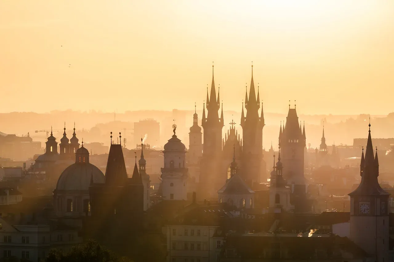 Dense Prague cityscape at golden hour, featuring numerous domes, spires, and historical towers against a hazy, orange sky.