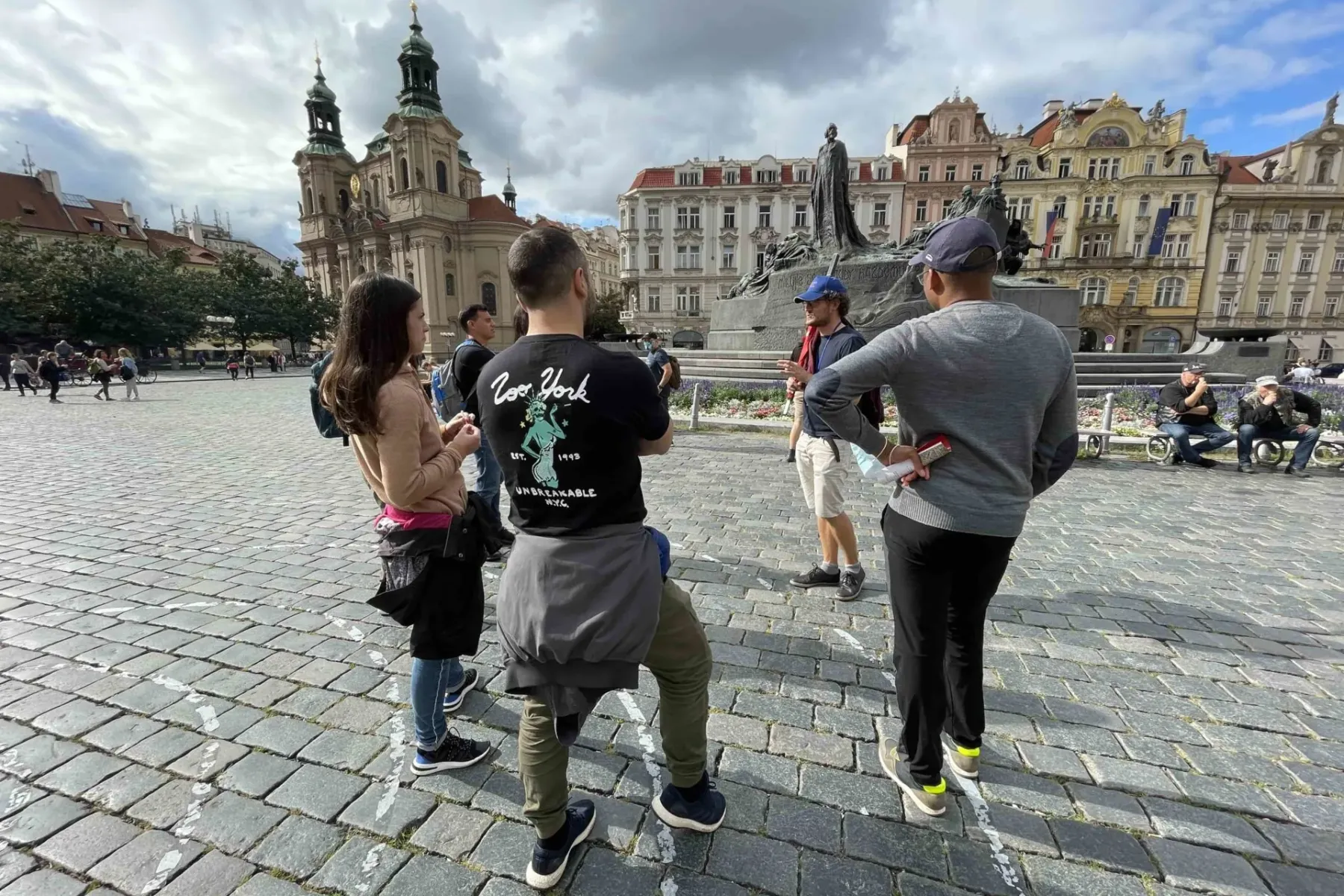 A small group of people listening to a guide in Prague's Old Town Square, with St. Nicholas Church and Jan Hus Monument.