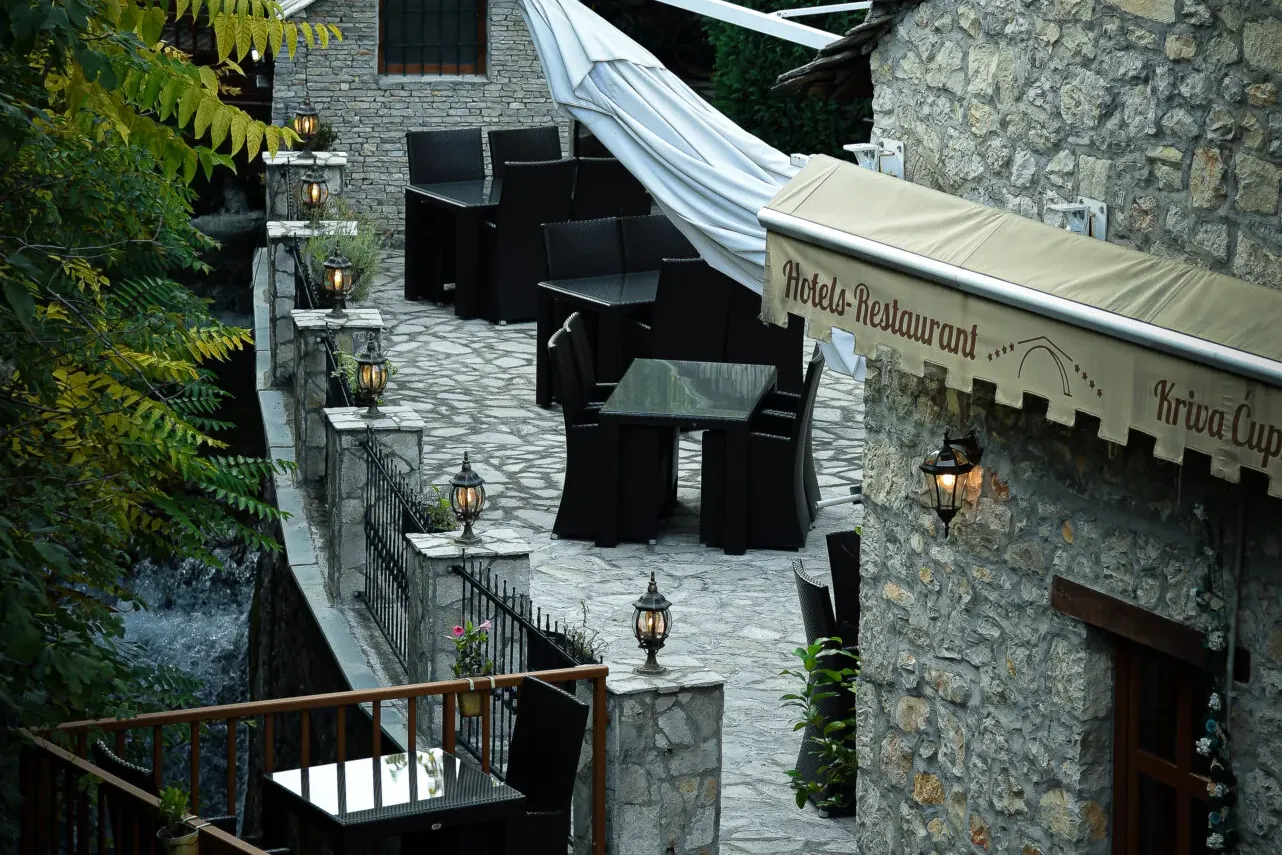 Outdoor patio of Hotel Kriva Ćuprija in Mostar, with stone walls, black wicker furniture, white awning, and a river flowin...