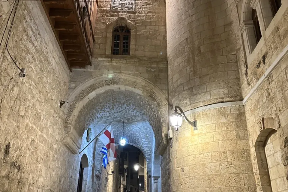 Narrow stone alley at night with an archway, steps, two motorbikes, and a building with a cross and wooden balcony.