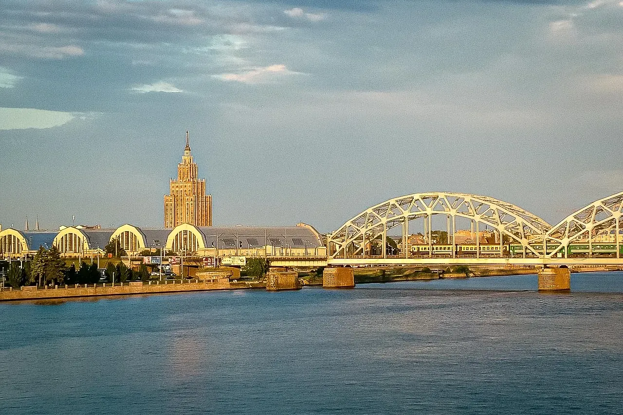 View across Daugava River in Riga showing a railway bridge with a green train, Central Market buildings, and the Latvian A...