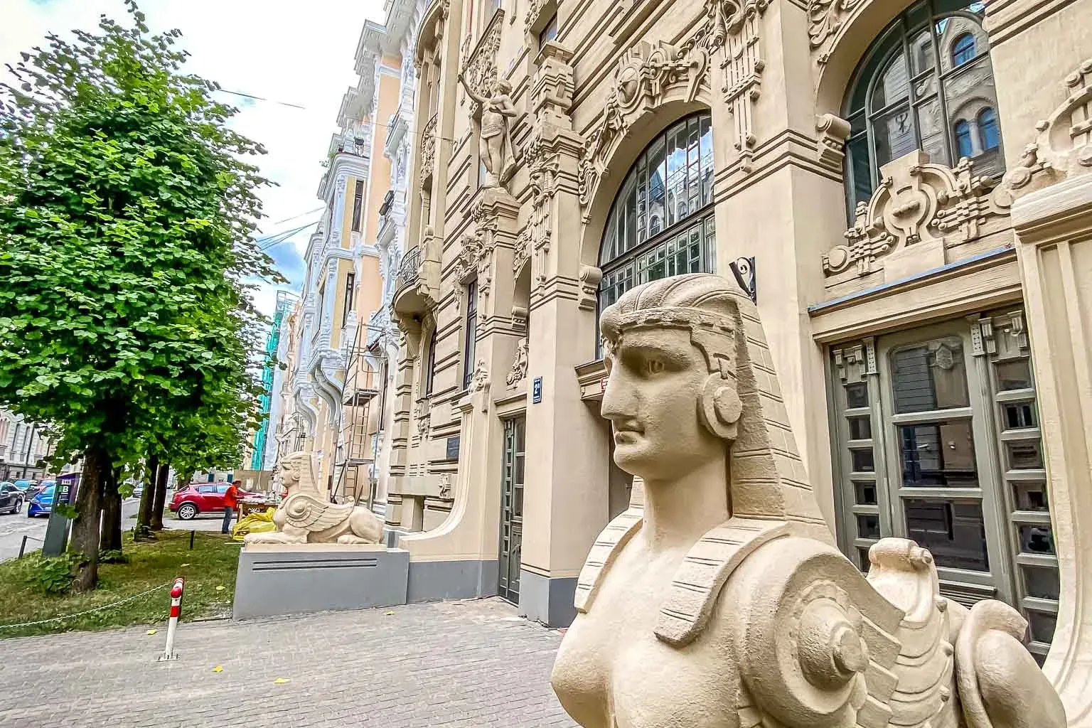 Light-colored sphinx statue with a human head and winged body on a plinth, in front of an ornate Art Nouveau building in R...