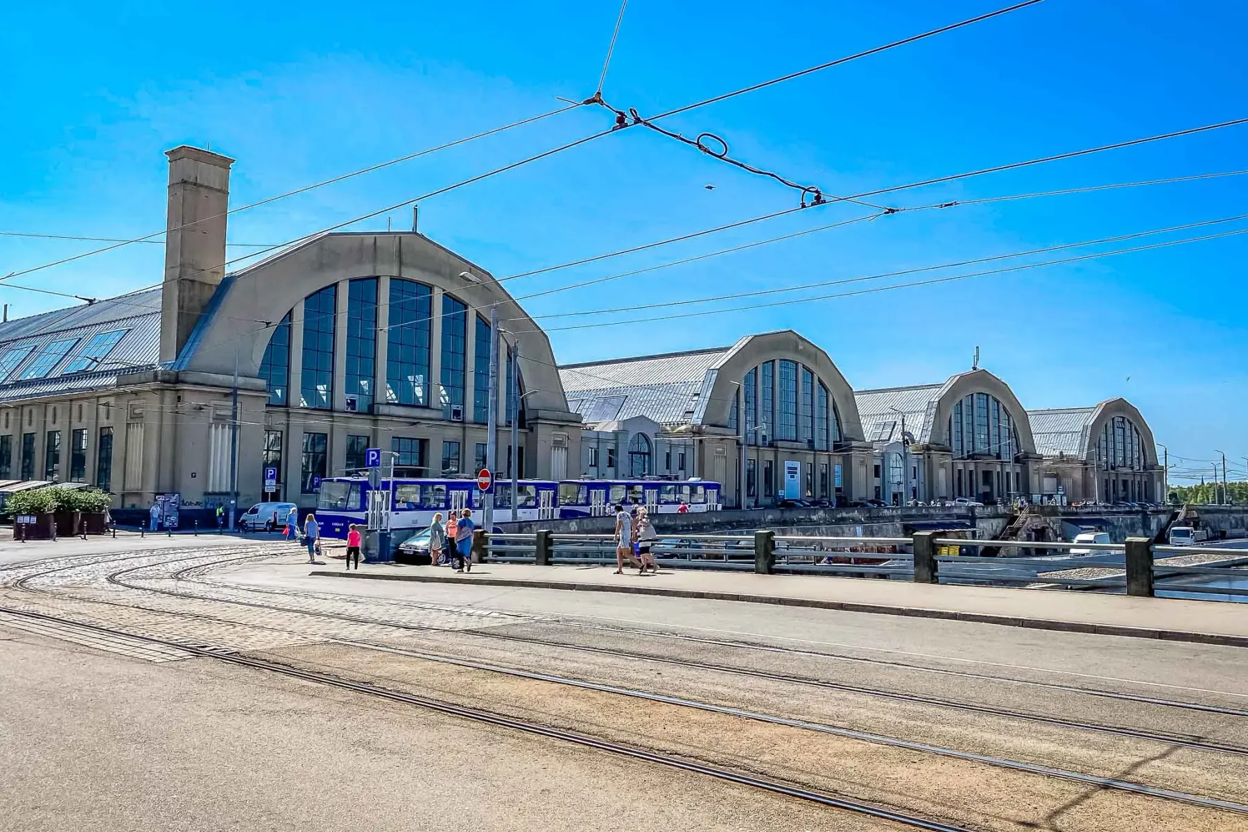 Five large market halls with arched roofs and windows, tram tracks in the foreground, people walking on a street in Riga.