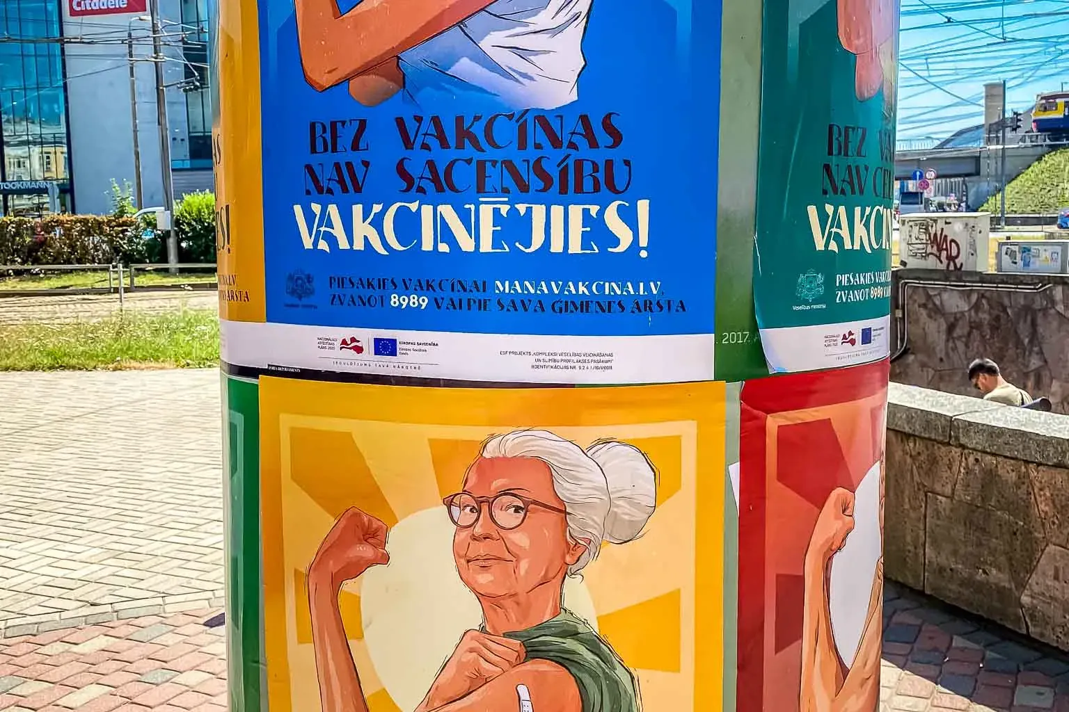 Cylindrical advertising pillar covered with posters for a vaccination campaign in Riga, featuring a woman playing tennis a...