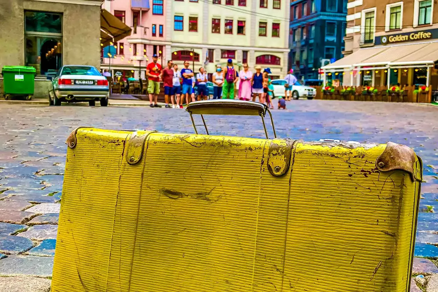 A bright yellow, old suitcase leaning on a colorful cobblestone street in Riga, with blurred historic buildings and people...