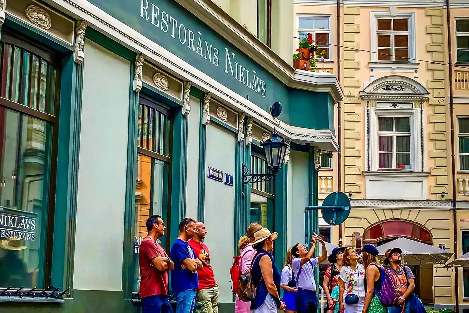 A group of people looking up at the facade of a light green building with "RESTORĀNS NIKLĀVS" sign. Historic buildings beh...