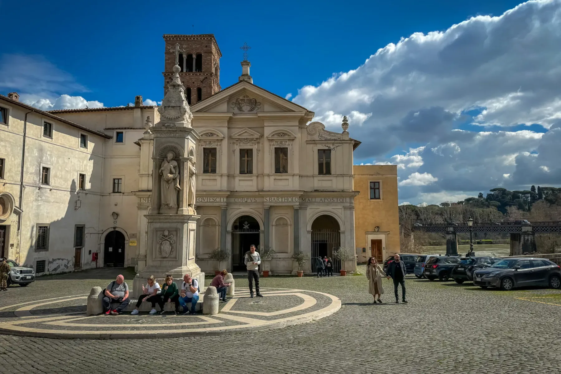 View of Basilica di San Bartolomeo and a monument on Tiber Island square, with people and cars under a cloudy blue sky in ...