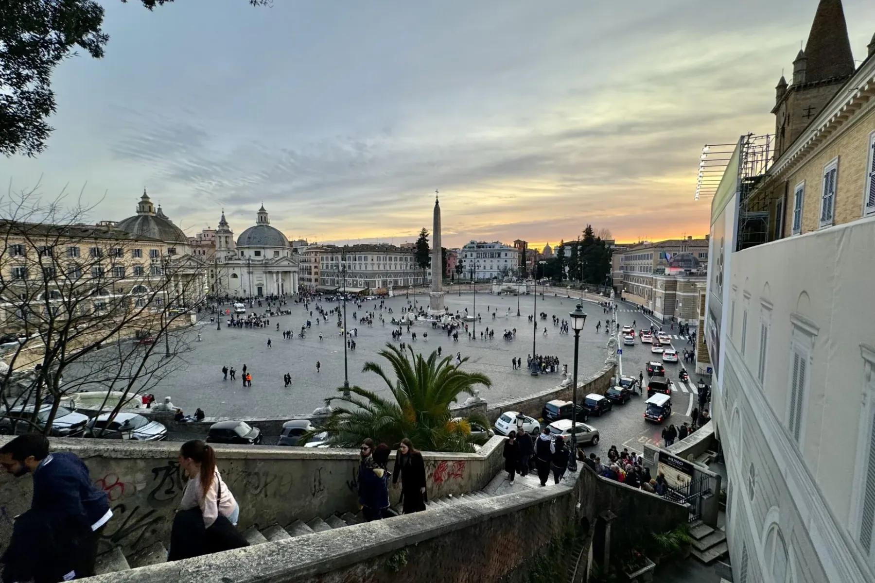 Elevated view of Piazza del Popolo in Rome at sunset, with an obelisk, two domed churches, crowds, and traffic.