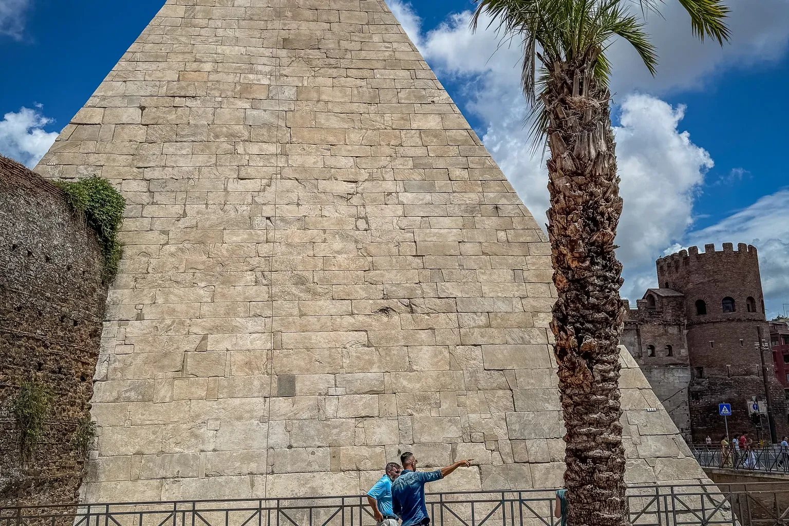 The Pyramid of Cestius in Rome, a large stone structure, with a tall palm tree and two tourists in front.