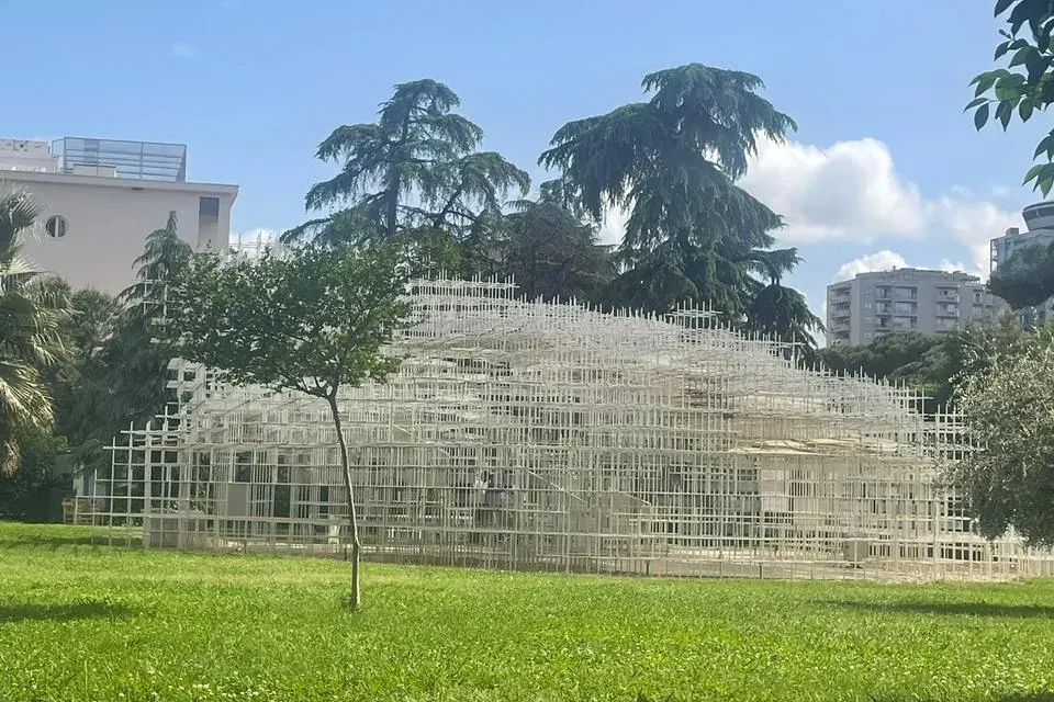Large white grid-like art installation in a green park under a blue sky with scattered clouds. Trees and buildings in the ...