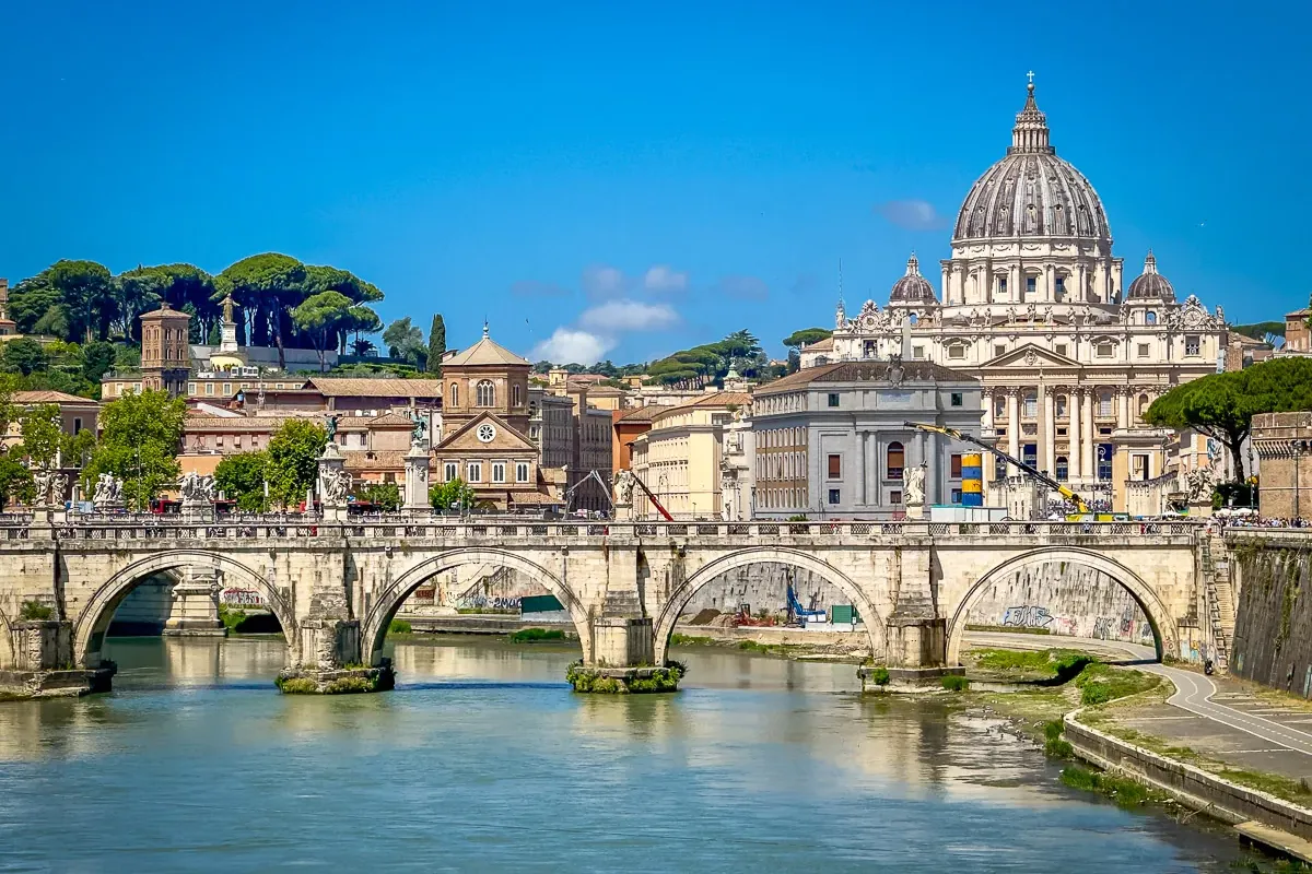 Sant'Angelo Bridge over the Tiber River, with St. Peter's Basilica dome and Roman buildings under a clear blue sky.