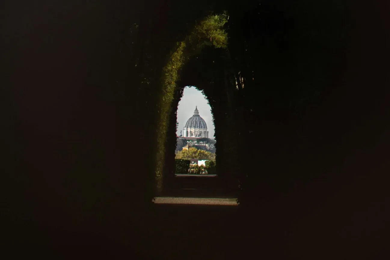 The dome of St. Peter's Basilica seen through a small, dark, keyhole-shaped opening in a hedge or wall with green foliage.