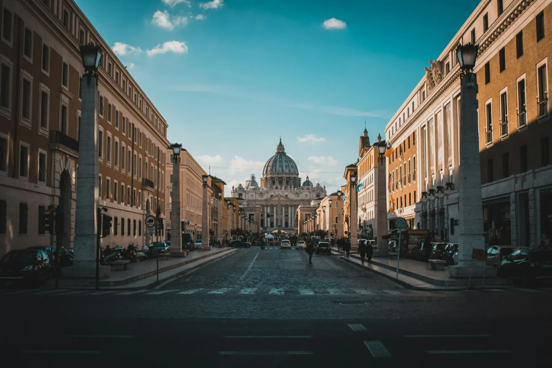 St. Peter's Basilica dome and facade viewed from a wide street lined with tall buildings and ornate street lamps under a b...