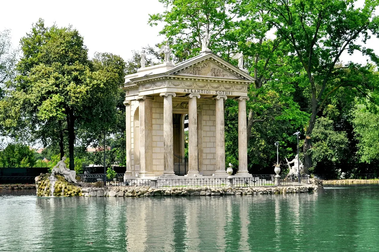 Classical temple on a small island in a serene pond, surrounded by lush green trees and sculptures, under a bright sky.