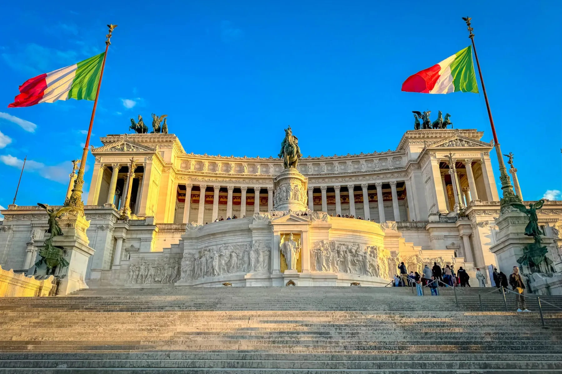 The Altar of the Fatherland in Rome, a white marble monument with columns, statues, and Italian flags, under a clear blue ...