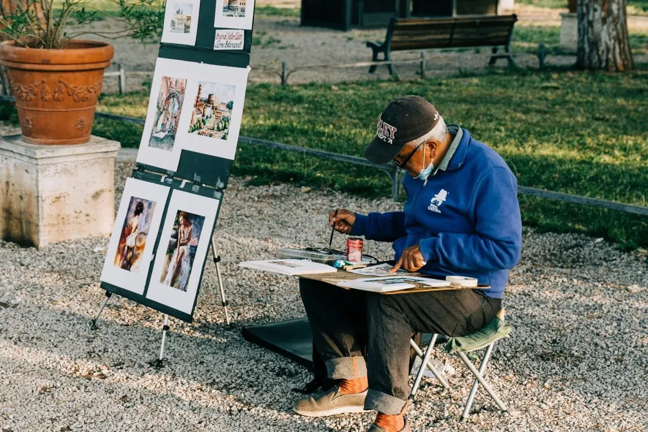 Artist painting outdoors on a gravel path next to an easel displaying watercolor paintings of city scenes. Trees and a ben...