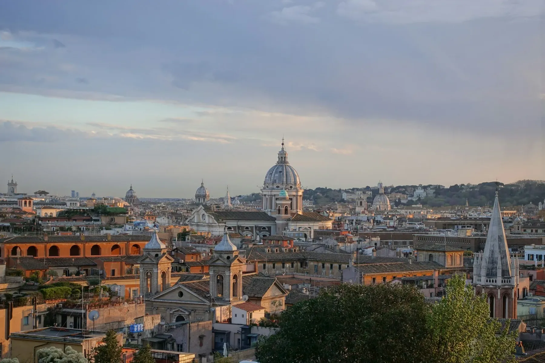 Panoramic view of Rome at sunset with multiple church domes and terracotta rooftops under a soft, cloudy sky.