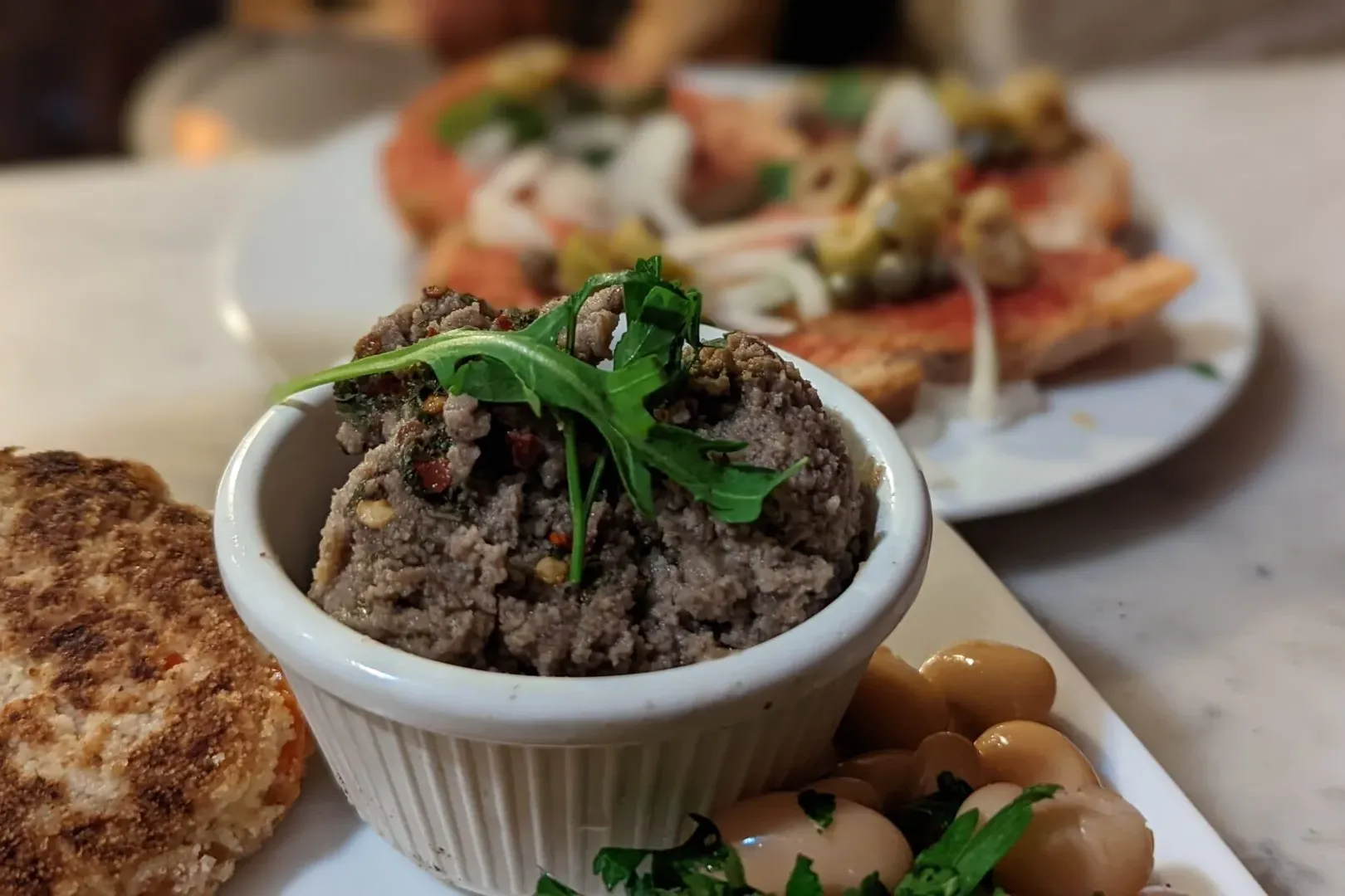 Close-up of a white ramekin with brown spread and arugula, a round fritter, and white beans with herbs. Blurred bruschetta...