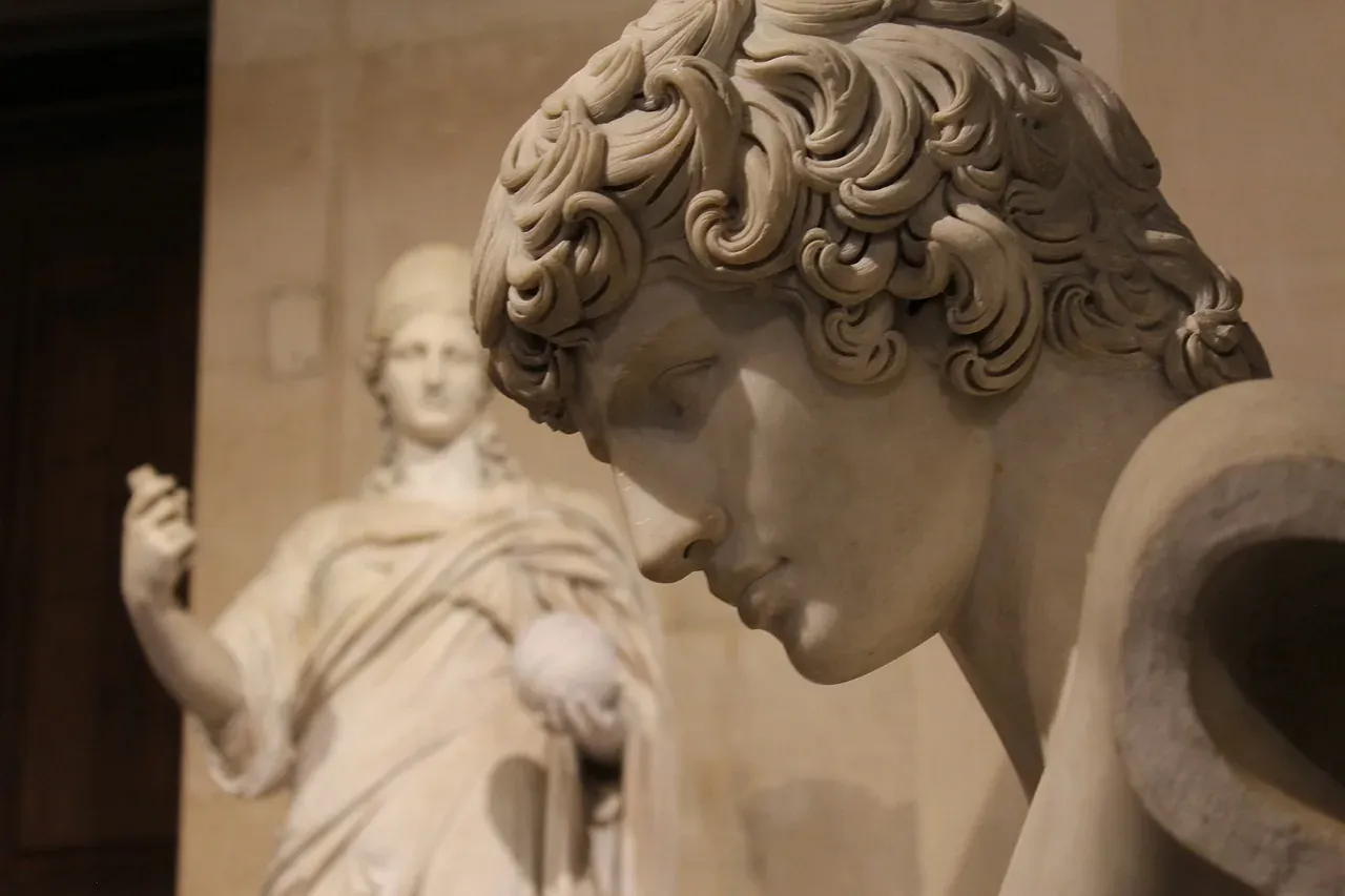 Close-up of a classical marble sculpture showing a person's head with curly hair, looking down. Another blurred statue is ...