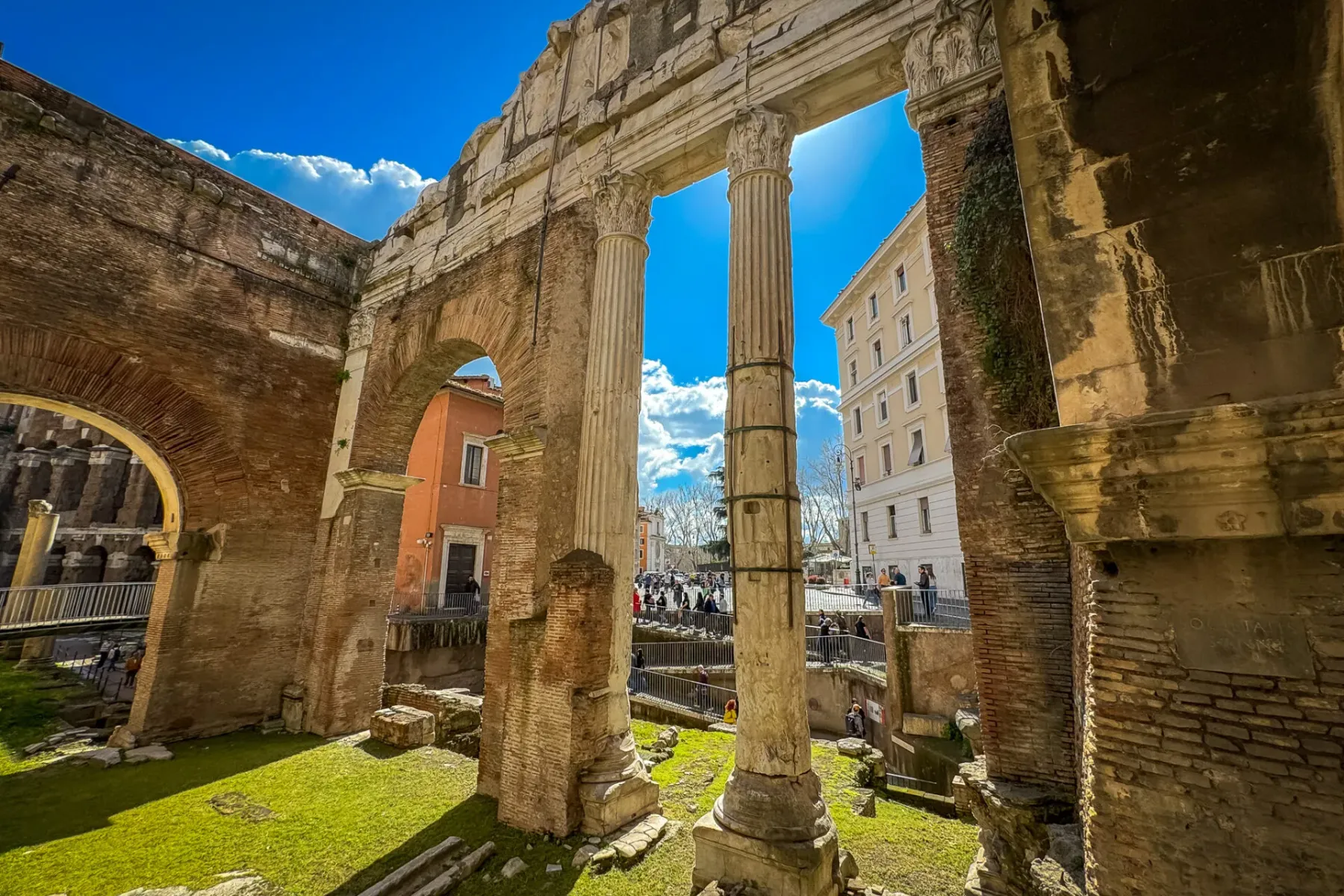 Ancient Roman brick ruins with arches and two tall stone columns. Green grass fills the excavated area, modern buildings v...