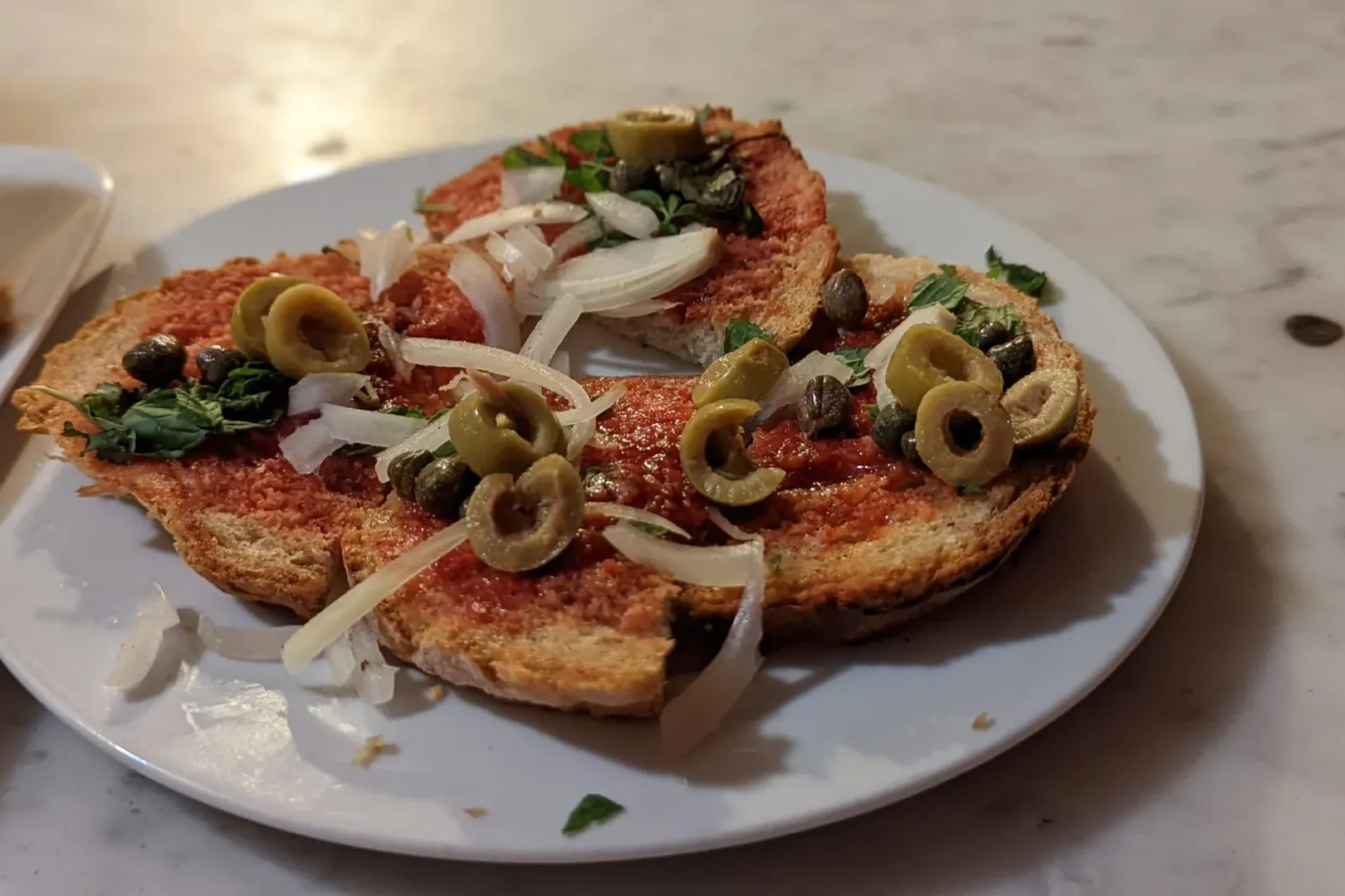 Toasted bread slices with red tomato spread, green olives, sliced white onion, capers, and herbs on a white plate.